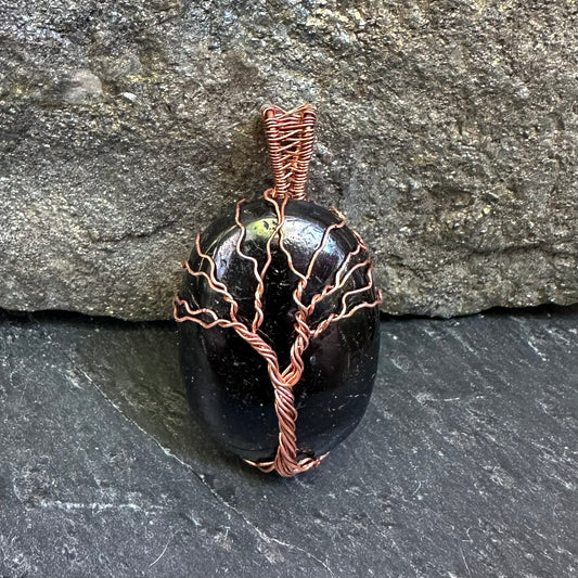 A polished black tourmaline stone pendant wrapped with copper wire in a tree-like pattern, set against a dark gray stone background.