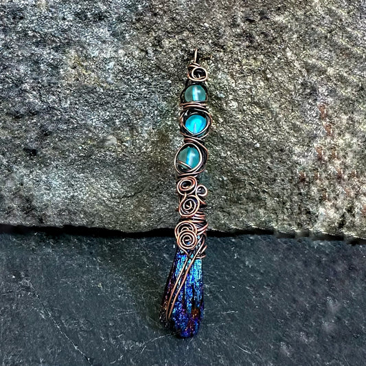 A handmade wire-wrapped pendant featuring three round turquoise-blue beads, copper swirls, and a textured blue and purple kyanite stone at the bottom, displayed against a dark gray stone background.