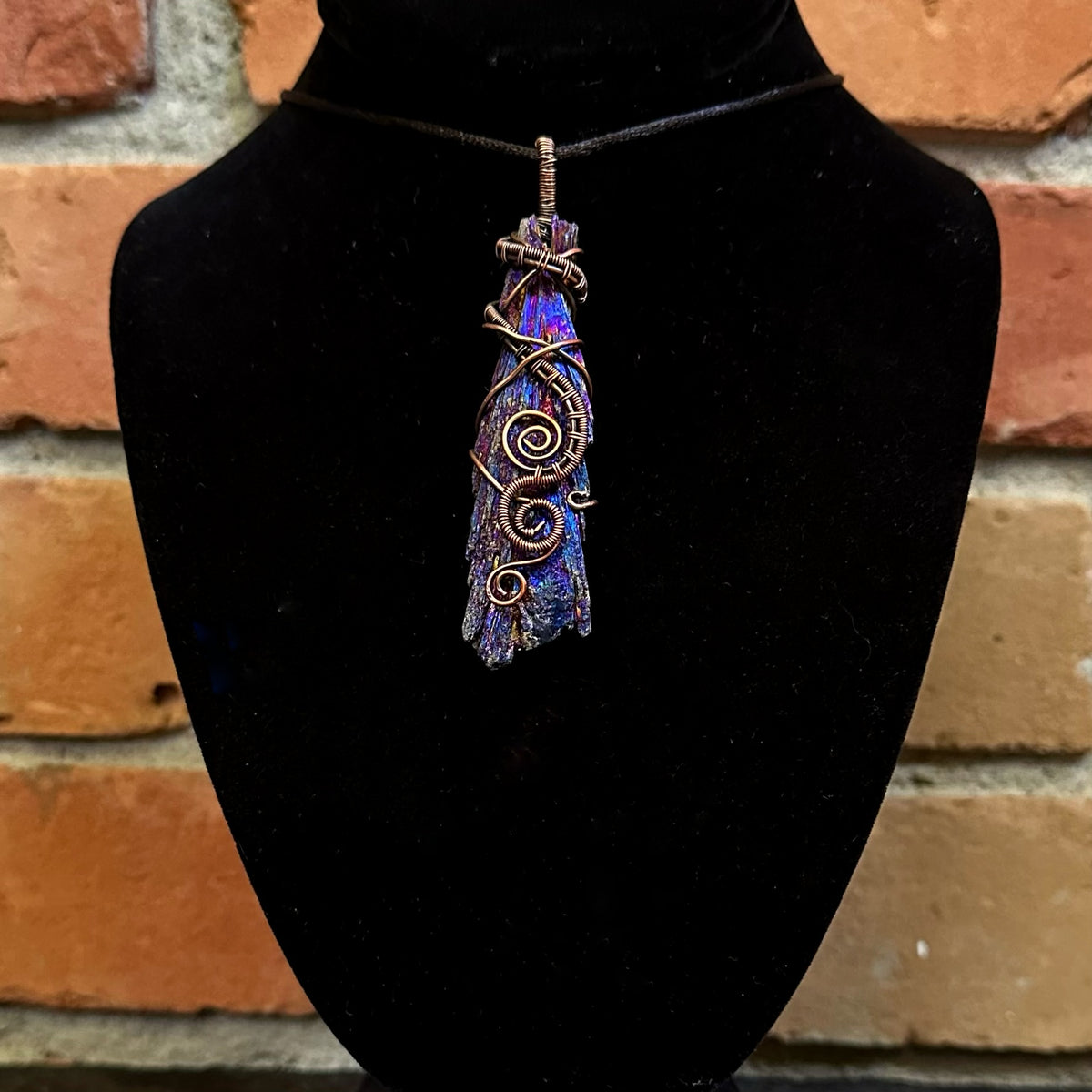 A handmade pendant necklace featuring a textured, iridescent blue and purple kyanite stone wrapped in copper wire, displayed on a black velvet bust against a rustic brick wall background.