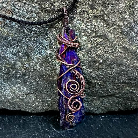 A handmade pendant necklace featuring a raw, jagged purple and blue iridescent kyanite stone wrapped with copper wire, hanging from a black cord against a textured gray rock background.