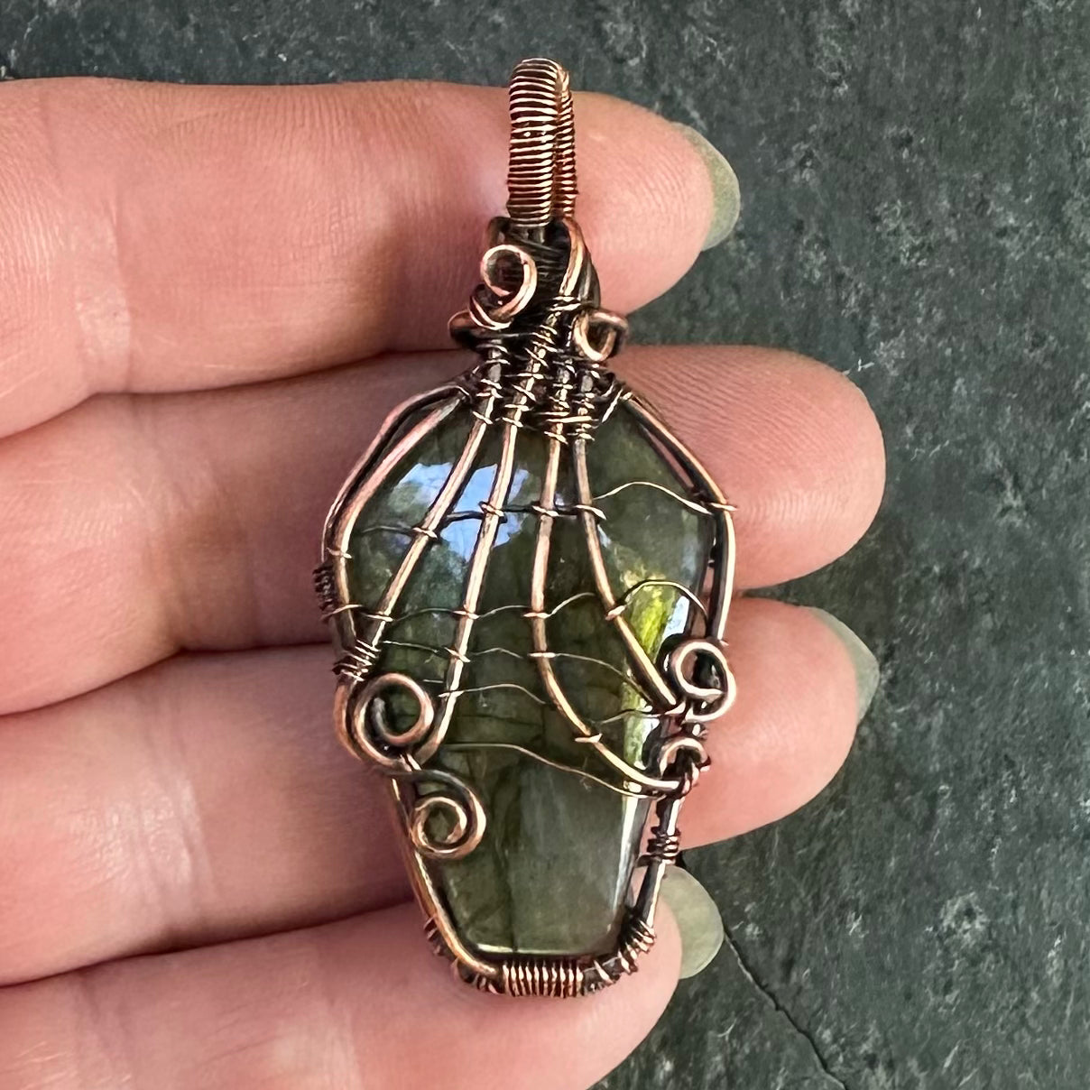 A handmade copper wire-wrapped pendant featuring a labradorite stone with a spider web pattern, held in a person's hand against a dark textured slate background.