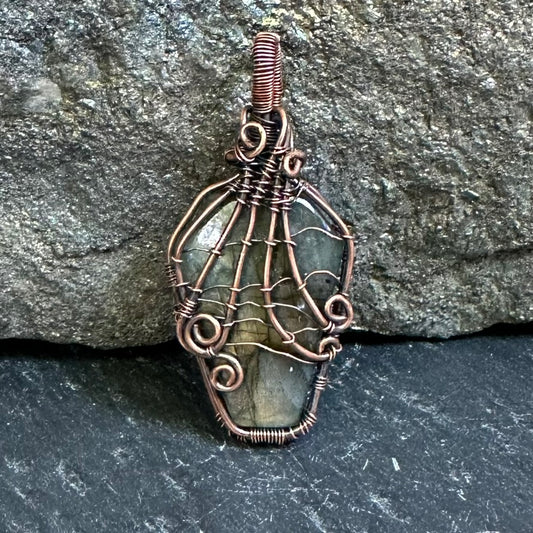 A handmade copper wire-wrapped pendant featuring a coffin-shaped natural labradorite stone with a spiderweb pattern, set against a dark textured rock background.