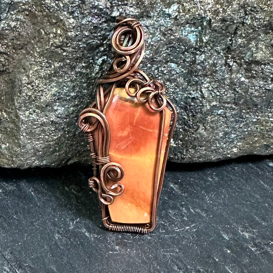 A coffin-shaped pendant featuring an orange and peach Maligano jasper stone inlaid with copper wire wrapping, displayed against a dark textured rock background.