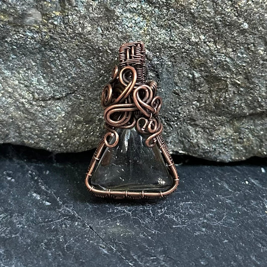 A handmade pendant featuring a shaman dream quartz wrapped in intricate copper wire, set against a dark textured stone background.