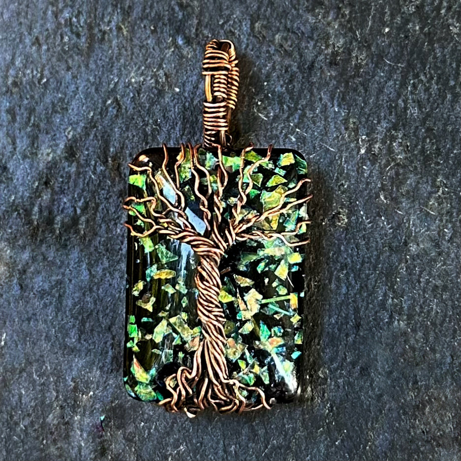 A rectangular pendant featuring a tree of life design wrapped in copper wire over a black and green iridescent mosaic background, shown on a dark textured stone surface.