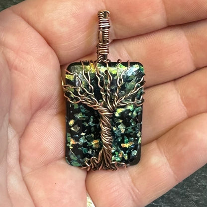 A rectangular pendant with a tree-of-life wire design over a dark green iridescent resin stone, held in a person's hand.