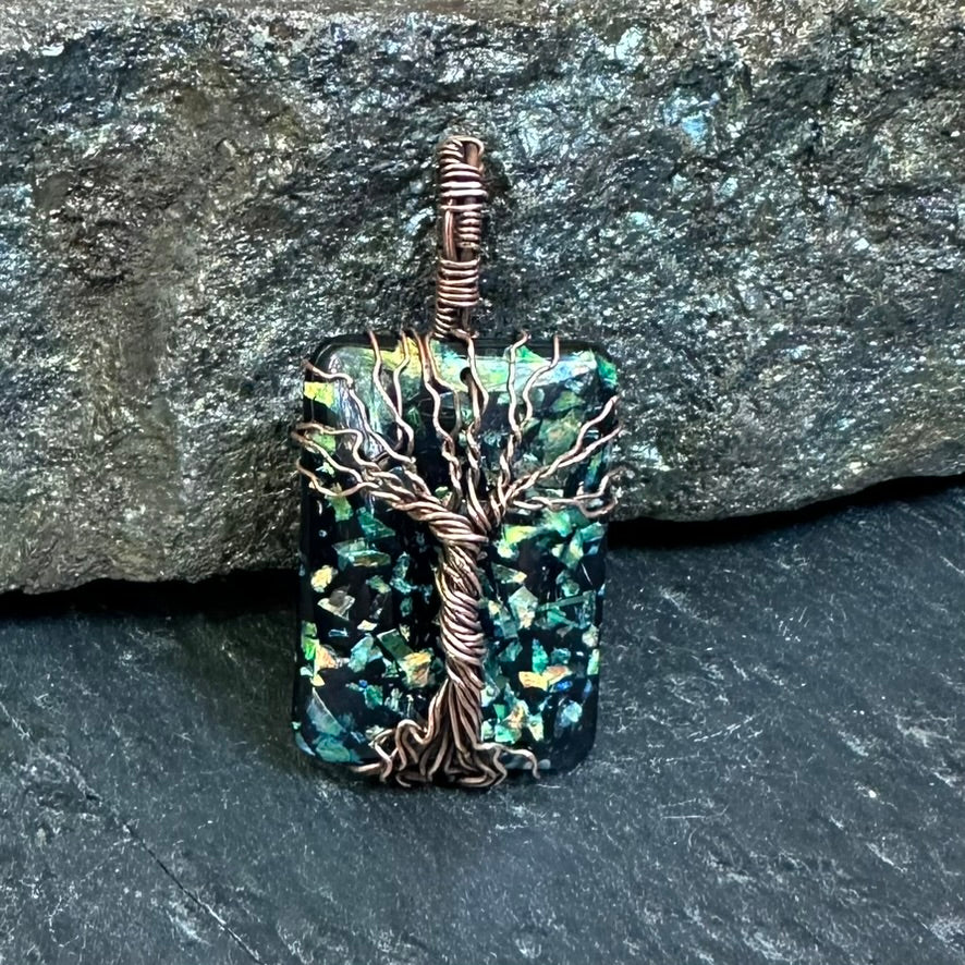 A handmade pendant featuring a square iridescent green and blue opal resin piece wrapped with a copper tree design, set against a dark textured stone background.