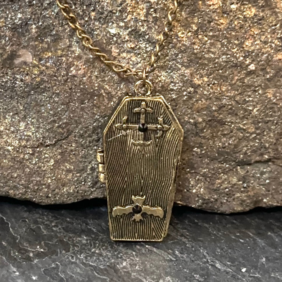 A coffin-shaped pendant necklace with a textured wood-grain finish, featuring a small cross on the door and a small dark gemstone accent, hanging from a gold-toned chain against a textured rock background.