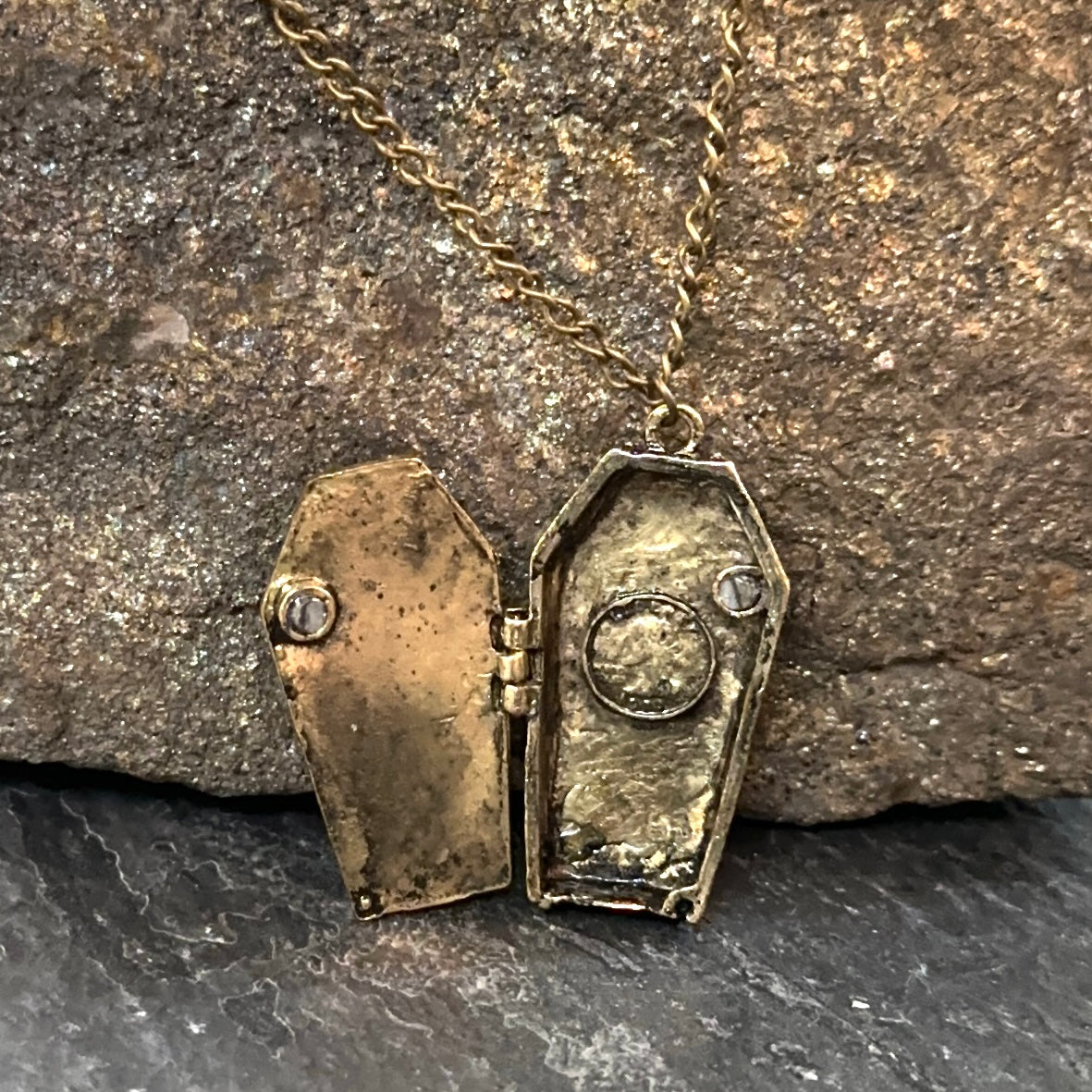 An antique-style gold coffin-shaped locket necklace, shown open with a small clear gemstone on each lid, resting on a textured brown rock surface.