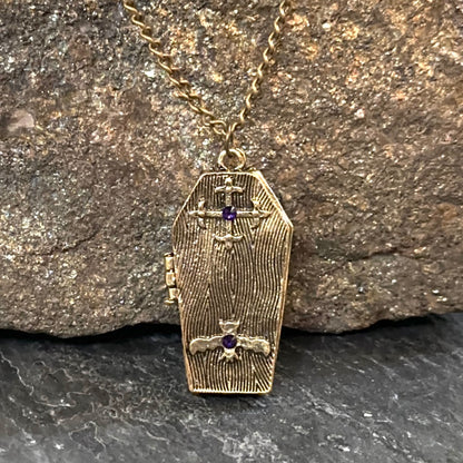An antique gold coffin-shaped pendant necklace with a textured wood-grain finish, featuring two small purple gemstone crosses, hanging from a gold chain against a textured brown rock background.