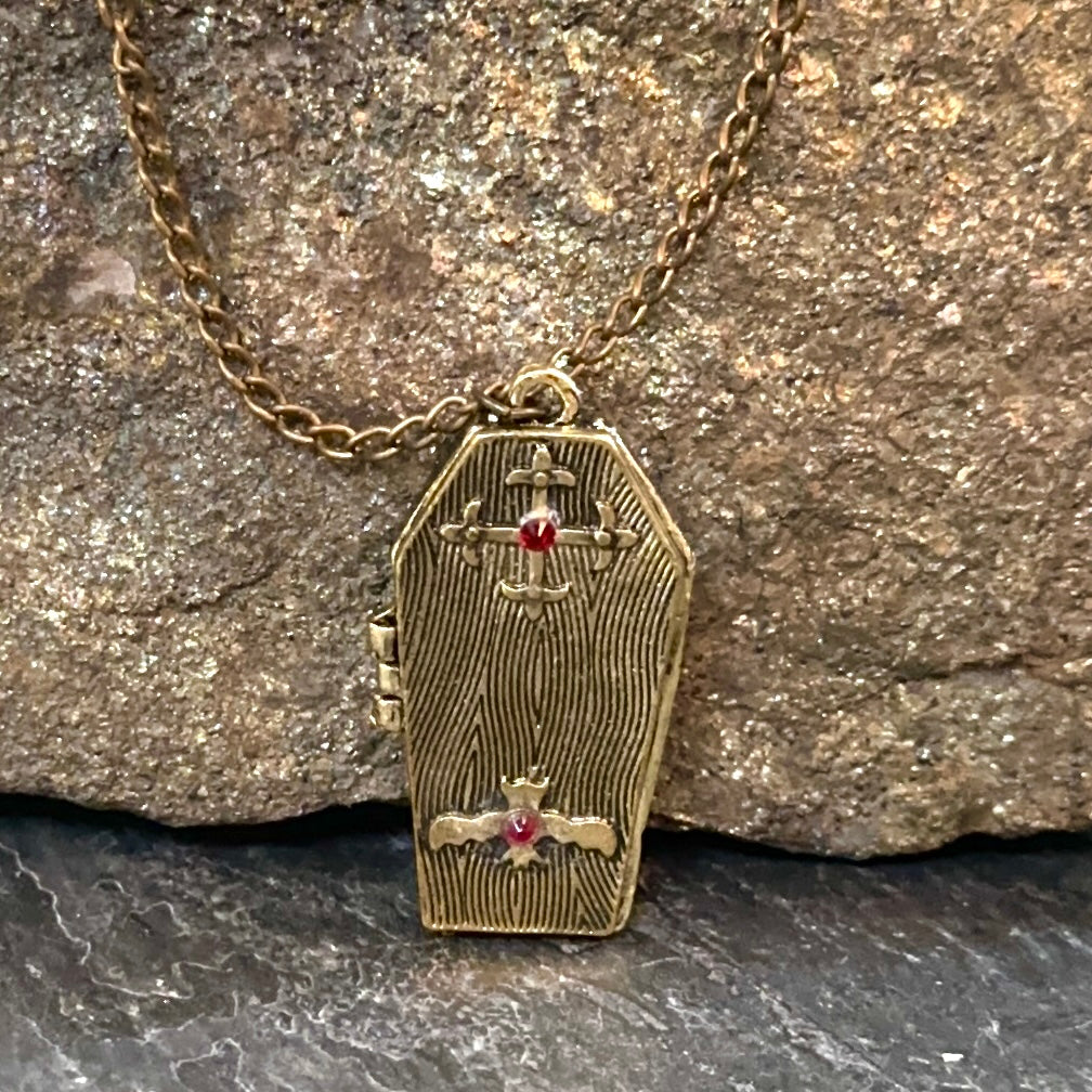 A coffin-shaped gold pendant necklace with engraved woodgrain details, a small cross at the top, a cross and dagger motif at the bottom, and small red gemstone accents, shown resting against a textured rock background.