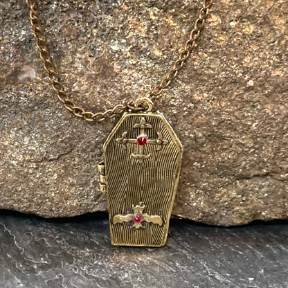 A coffin-shaped gold pendant necklace with engraved woodgrain details, a small cross at the top, a cross and dagger motif at the bottom, and small red gemstone accents, shown resting against a textured rock background.