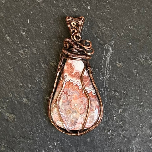A teardrop-shaped wire-wrapped pendant featuring a crazy lace agate stone, set in a copper-toned metal frame, displayed on a dark gray slate surface.