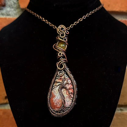 A teardrop-shaped pendant necklace with a crazy lace agate stone featuring a copper wire wrapped tree of life design, displayed on a black velvet jewelry bust.