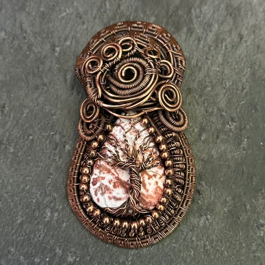 A copper-wrapped wire art piece featuring a tree of life design over a crazy lace agate stone, displayed on a dark gray slate surface.