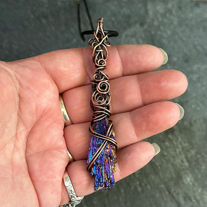 A hand holding a copper-wrapped pendant featuring a blue and purple iridescent kyanite stone, set against a dark textured background.
