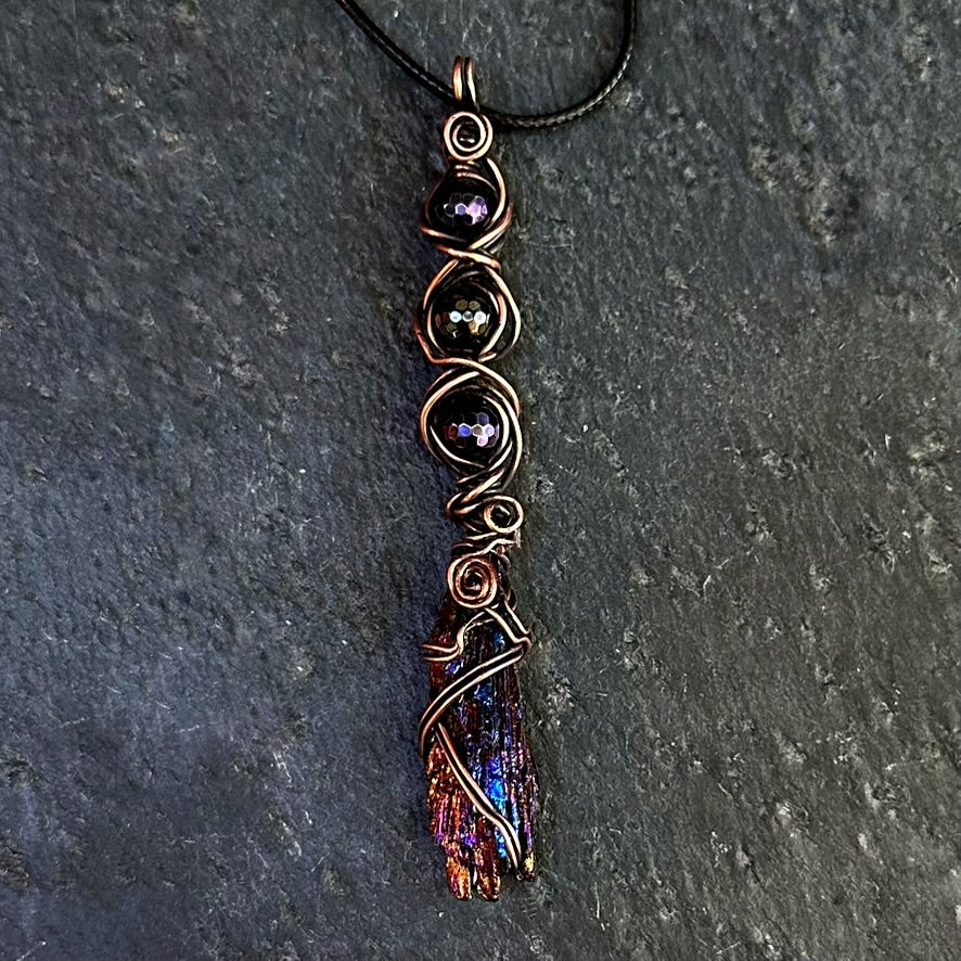 A handmade wire-wrapped pendant necklace featuring a dark iridescent kyanite stone with copper-toned wrapping and dark gemstone beads, displayed on a dark gray slate surface.