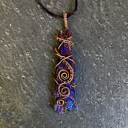 A handmade copper wire-wrapped iridescent purple and blue kyanite stone on a black cord, displayed on a textured gray slate surface.