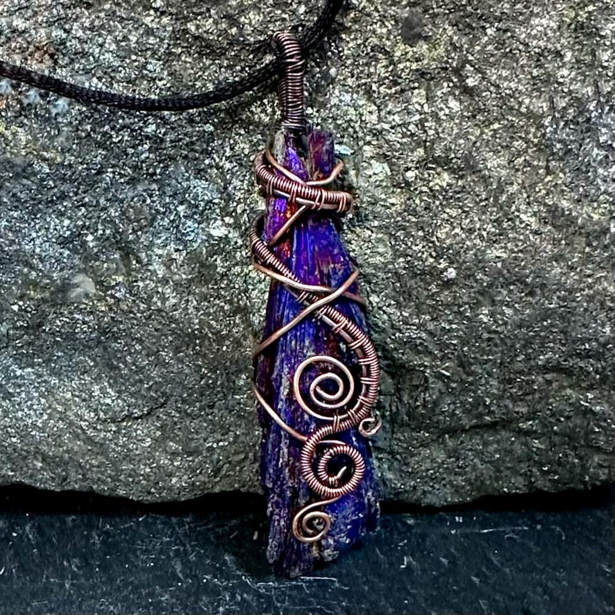 A handmade pendant necklace featuring a raw, jagged purple and blue iridescent kyanite stone wrapped with copper wire, hanging from a black cord against a textured gray rock background.