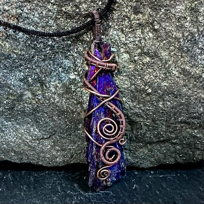 A handmade pendant necklace featuring a raw, jagged purple and blue iridescent kyanite stone wrapped with copper wire, hanging from a black cord against a textured gray rock background.
