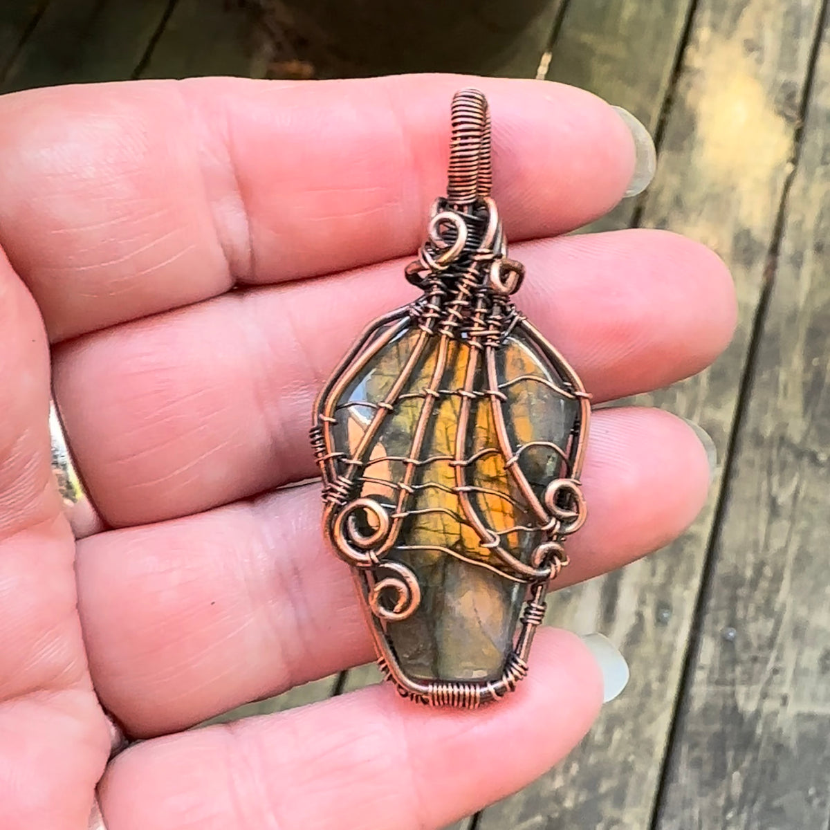 A handmade wire-wrapped pendant featuring a coffin shaped labradorite stone, wrapped with a spiderweb design, held in a person's hand against a blurred wooden background.
