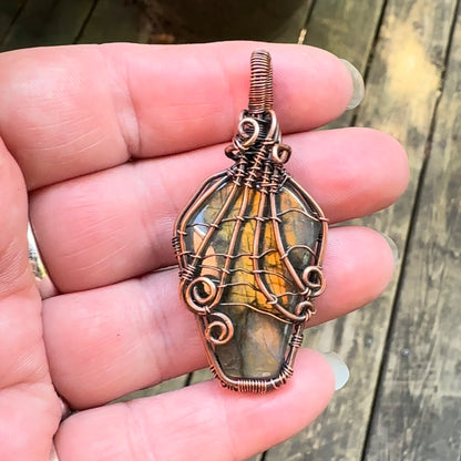 A handmade wire-wrapped pendant featuring a coffin shaped labradorite stone, wrapped with a spiderweb design, held in a person's hand against a blurred wooden background.