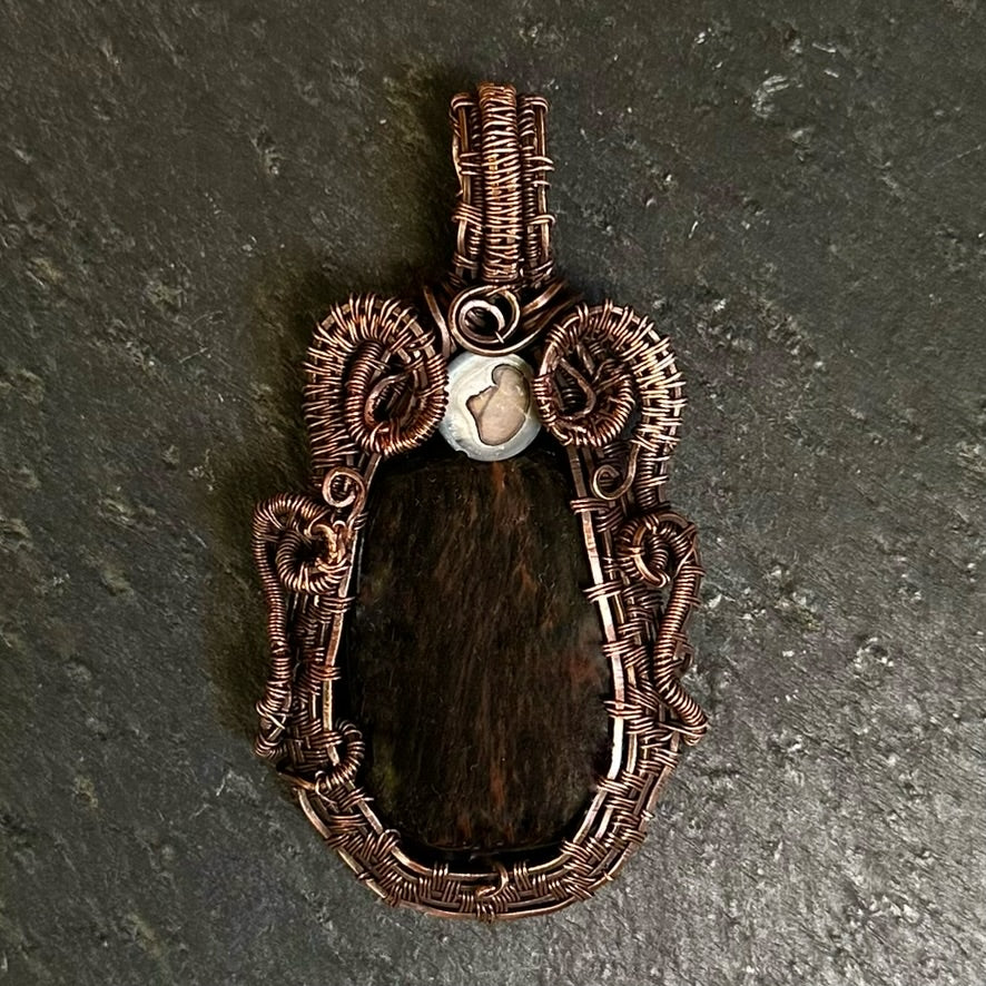 A handmade copper wire pendant with a rustic, organic coiled design and a central mahogany stone, displayed on a dark textured slate surface.