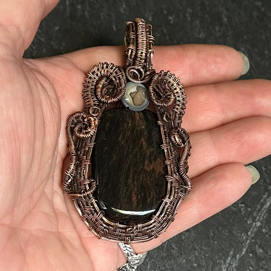 A handmade pendant featuring a mahogany stone inlay surrounded by intricate copper wire wrapping, held in a person's hand against a dark textured background.