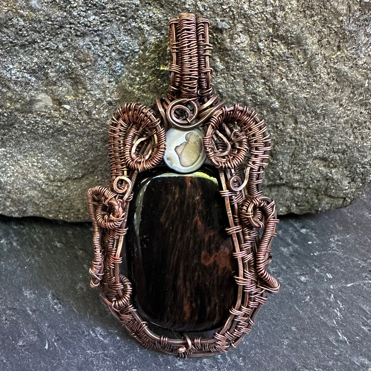 A handmade wire-wrapped pendant featuring a mahogany stone centerpiece, a small white stone, and intricate copper-toned wrapping, displayed against a gray stone background.
