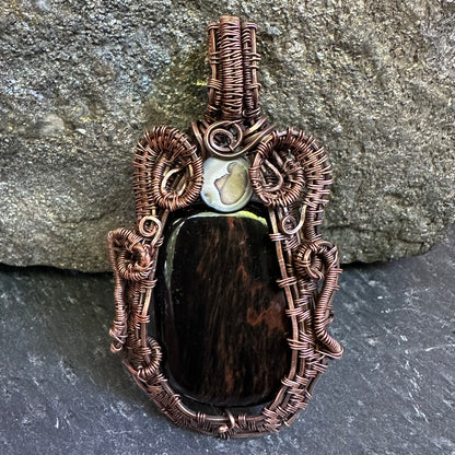 A handmade wire-wrapped pendant featuring a mahogany stone centerpiece, a small white stone, and intricate copper-toned wrapping, displayed against a gray stone background.
