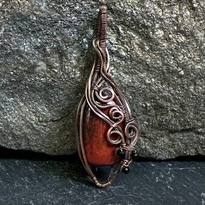 A copper wire-wrapped pendant featuring a polished red jasper stone, displayed against a textured gray rock background.