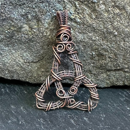 A copper-wrapped wire pendant shaped like a stylized triquetra or Celtic knot, displayed against a textured gray rock background.